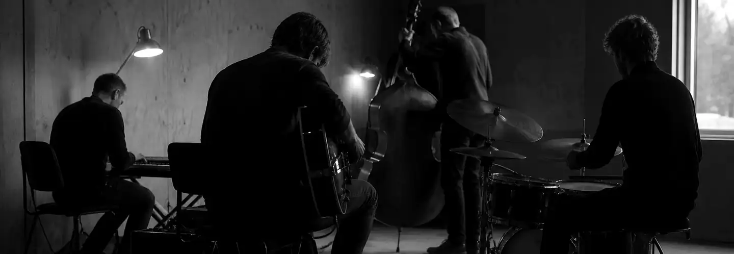 Fire musikere øver i et dunkelt øvelokale med kontrabas, trommesæt, guitar og keyboard.